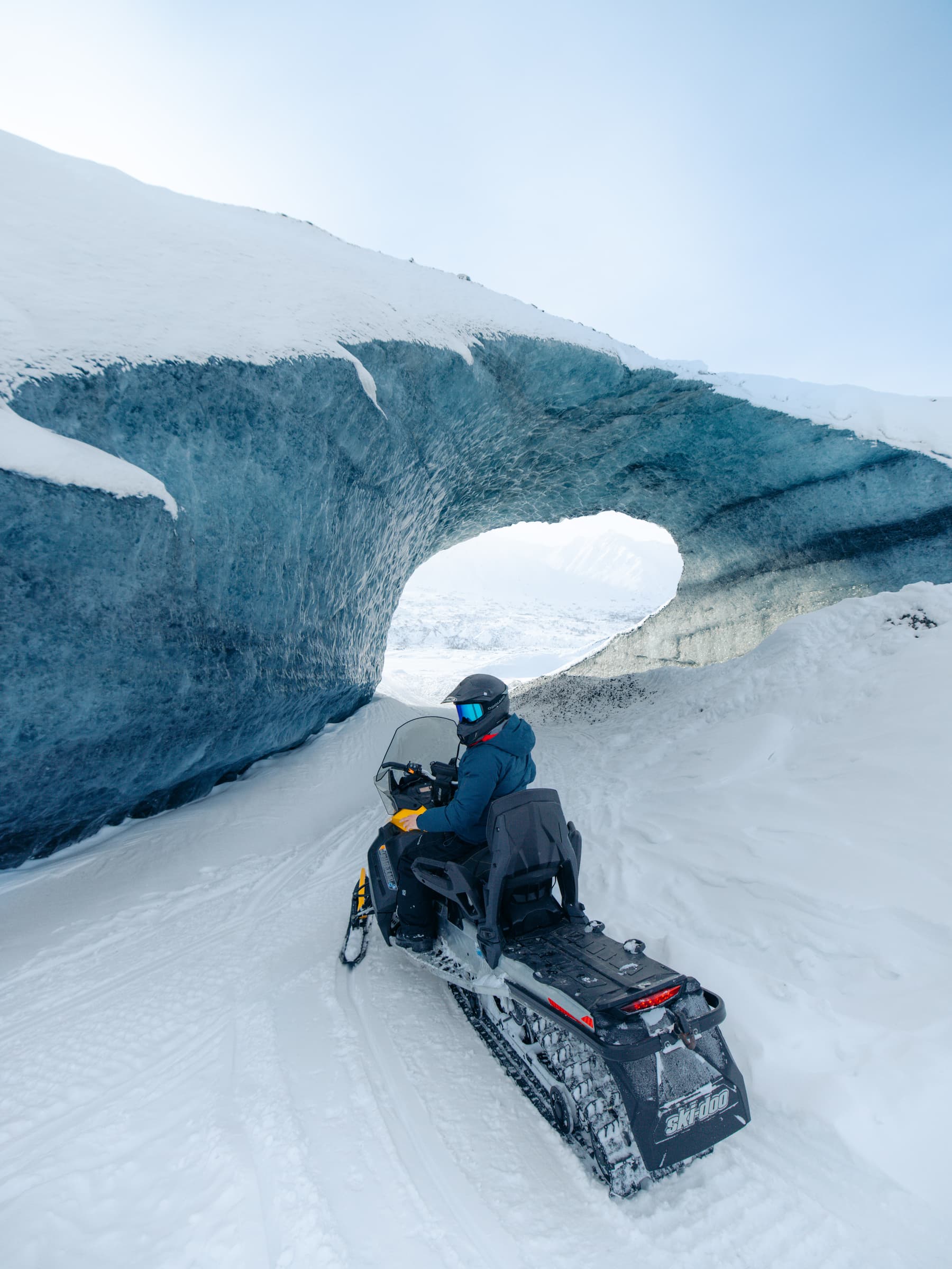 A guided snowmobile rider beneath a blue glacial ice arch in the Alaska backcountry.